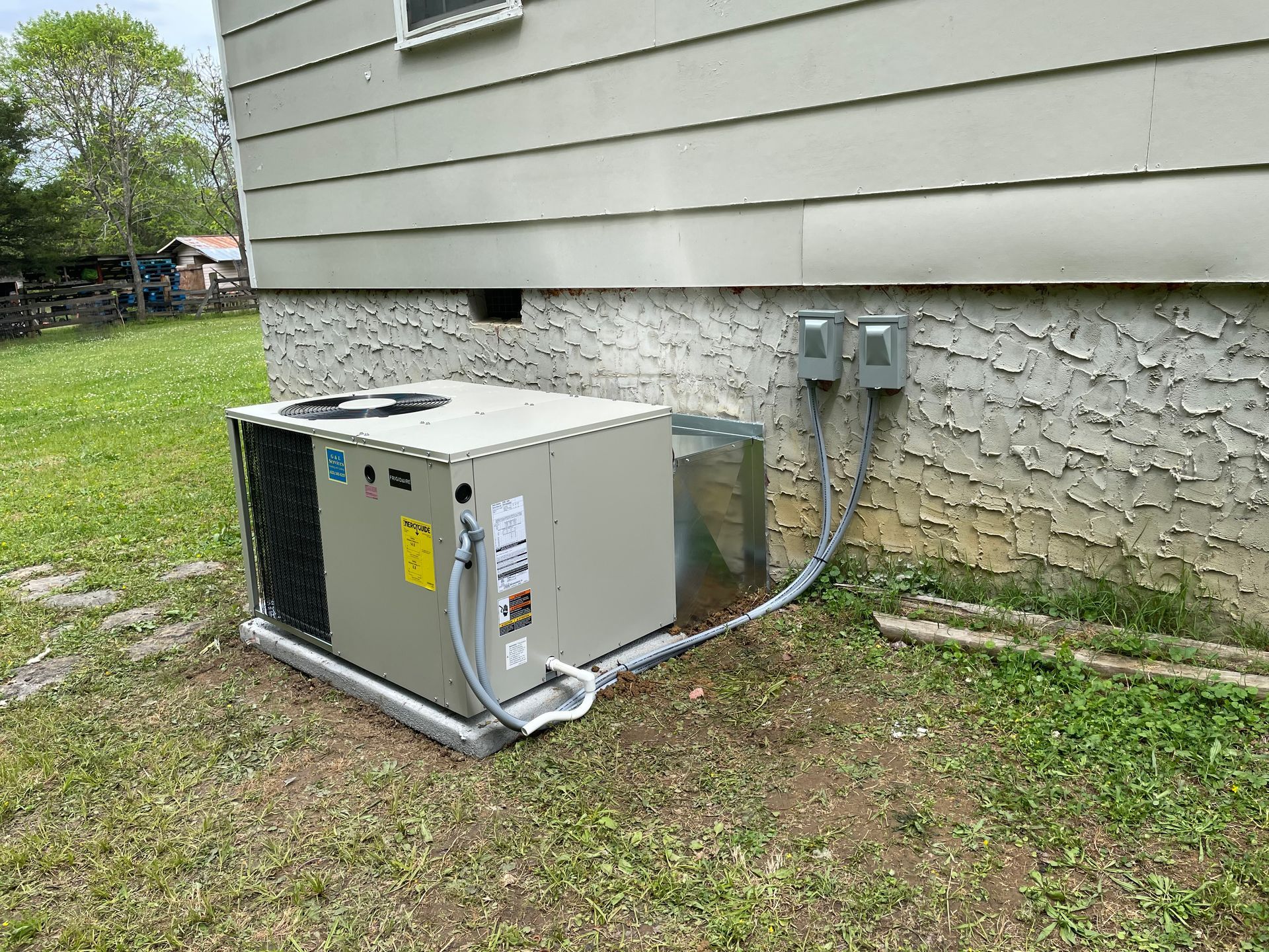 Outdoor central air conditioner unit beside a house, with electrical boxes and conduit on the exterior wall.