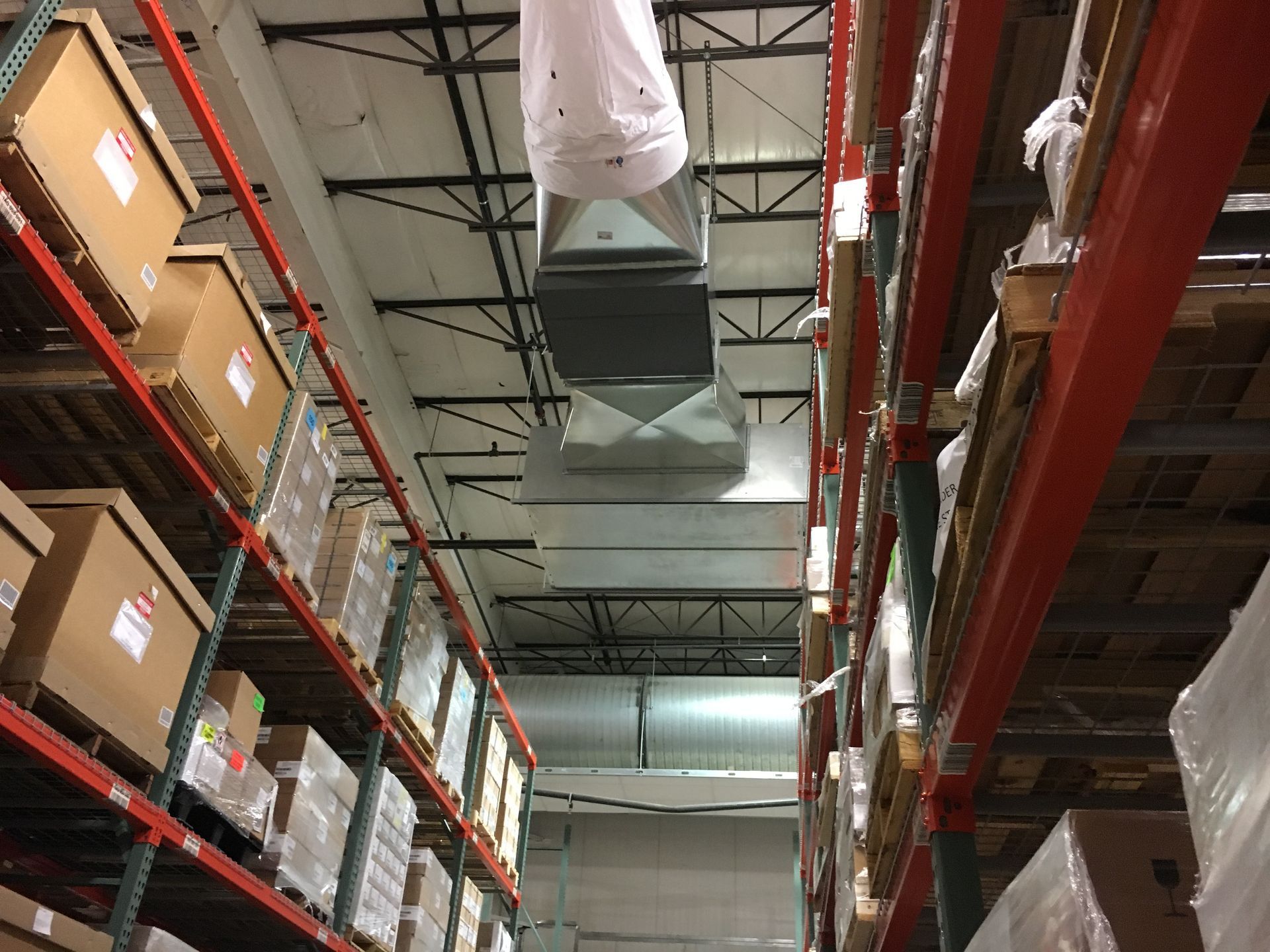 Warehouse aisle with tall shelving, stacked boxes, and large ceiling ducts overhead