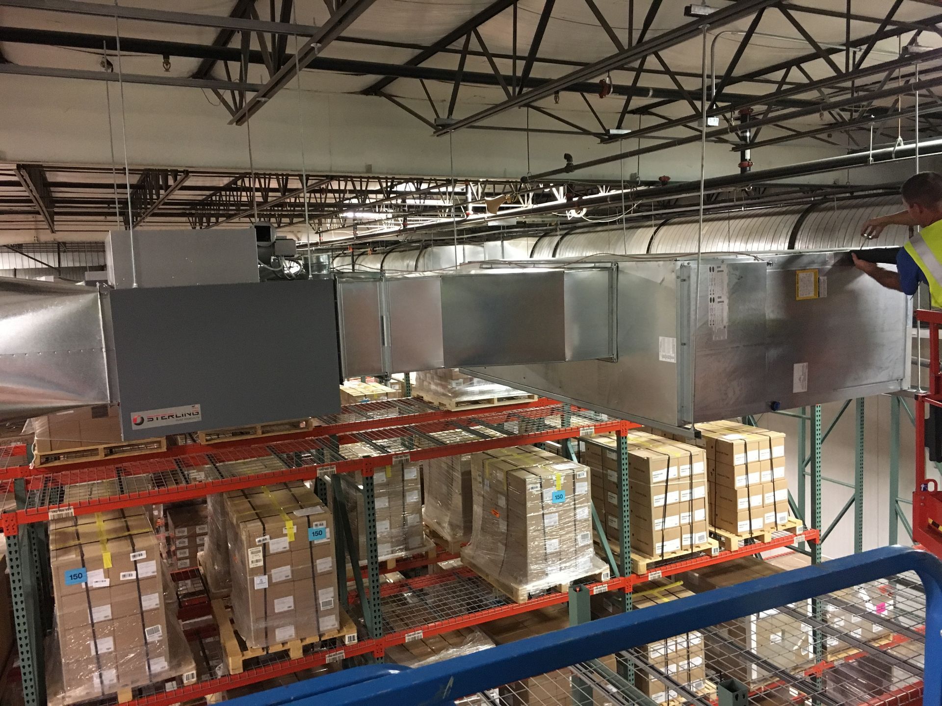 Warehouse interior with stacked boxes, metal racks, and ductwork beneath the ceiling