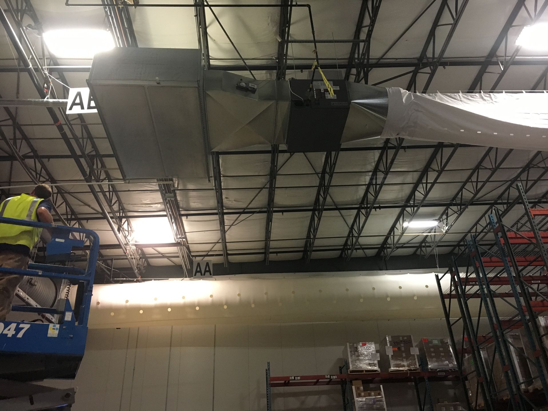 Warehouse workers installing large ceiling ductwork on a scaffold in an industrial interior