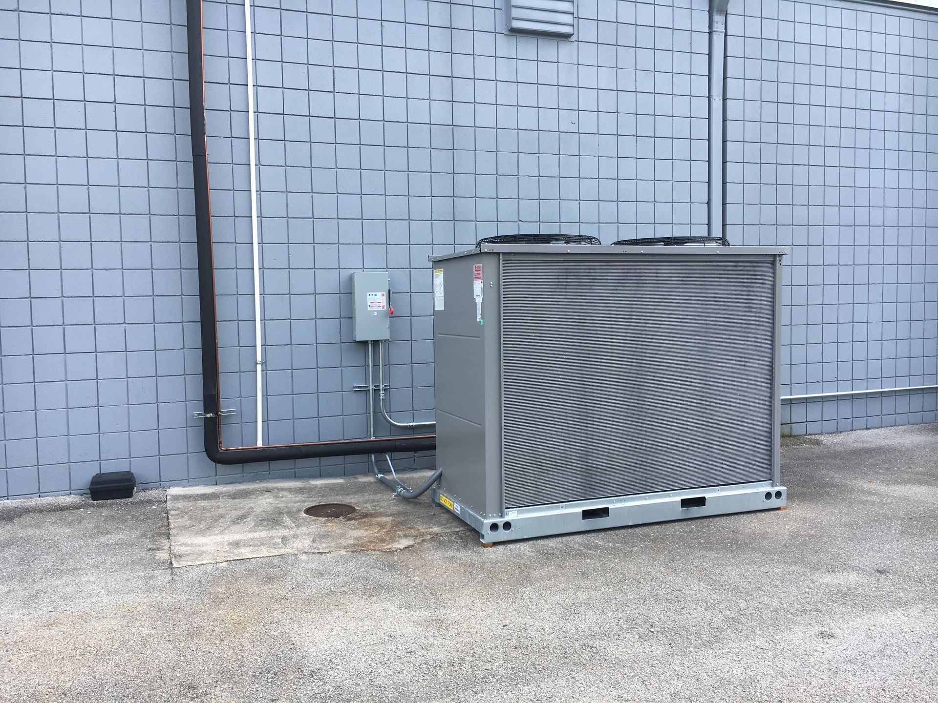 Outdoor HVAC unit beside a gray building wall with utility pipes and meters.
