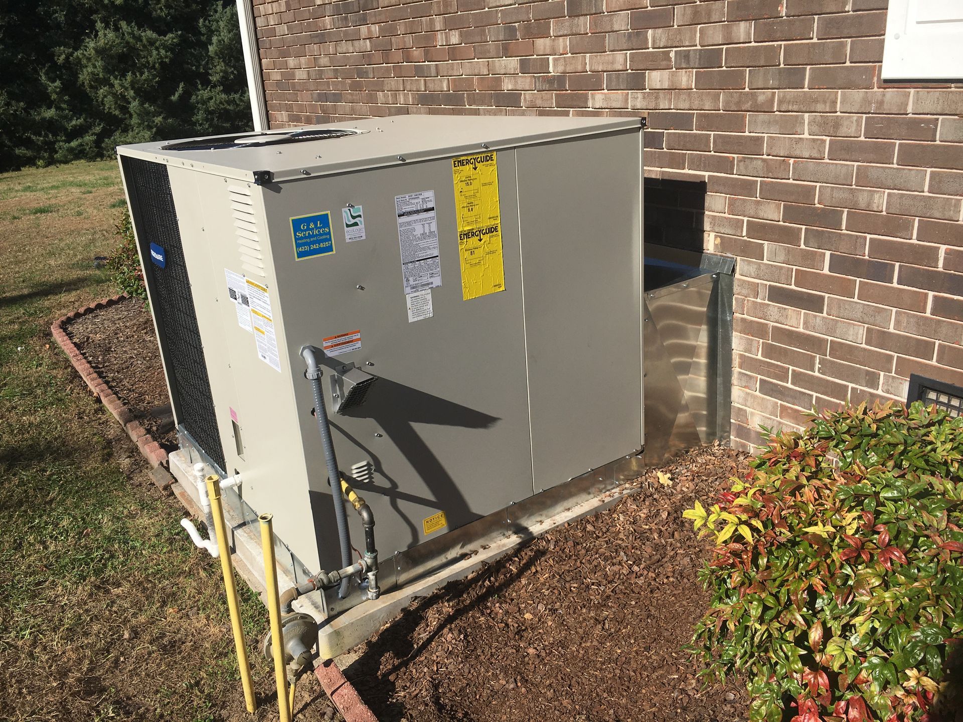 Outdoor HVAC unit beside a brick house wall, with pipes and a yellow warning label in a landscaped yard