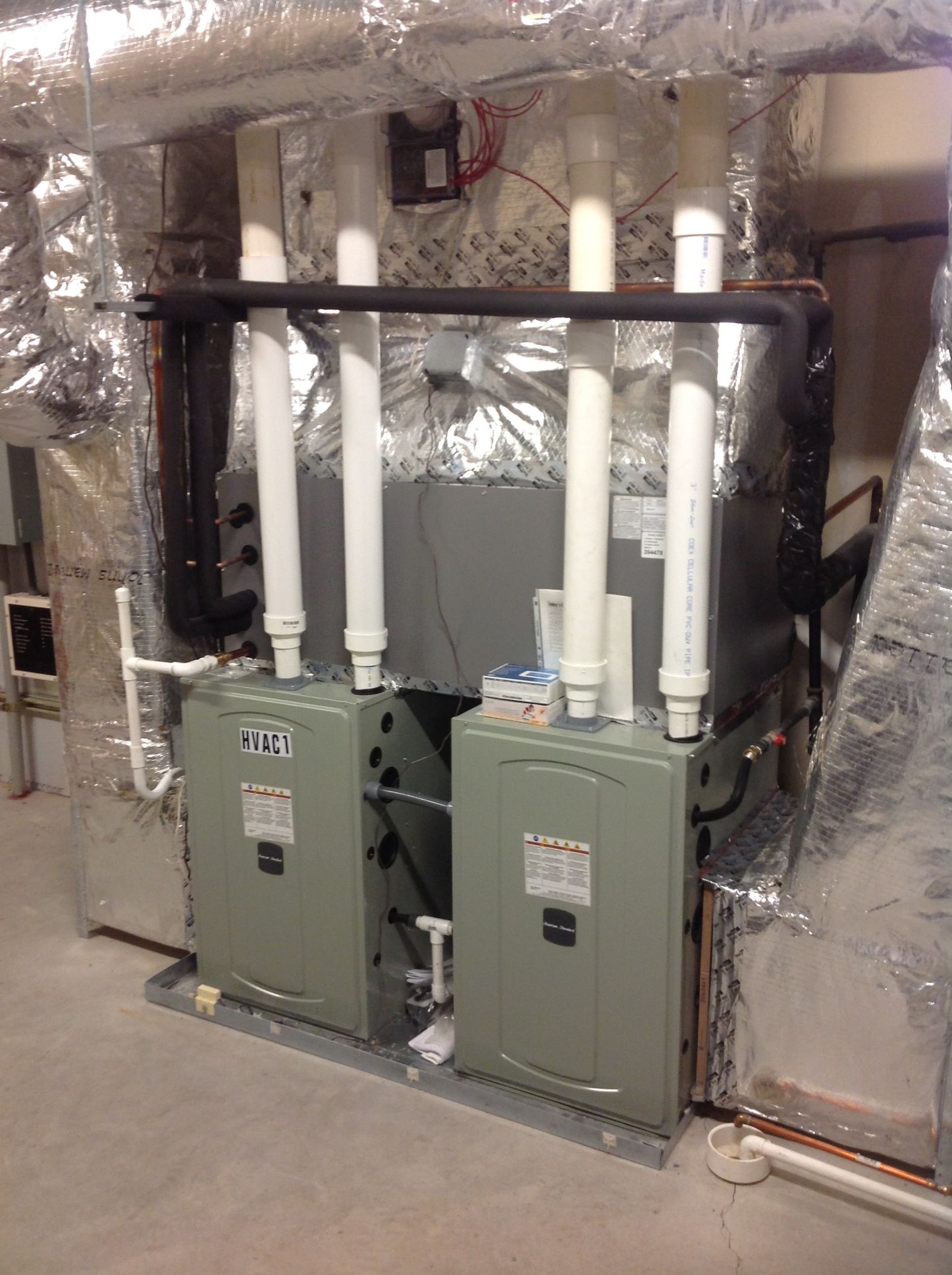 Two green furnaces with white PVC vent pipes in a utility room, surrounded by insulated ductwork.