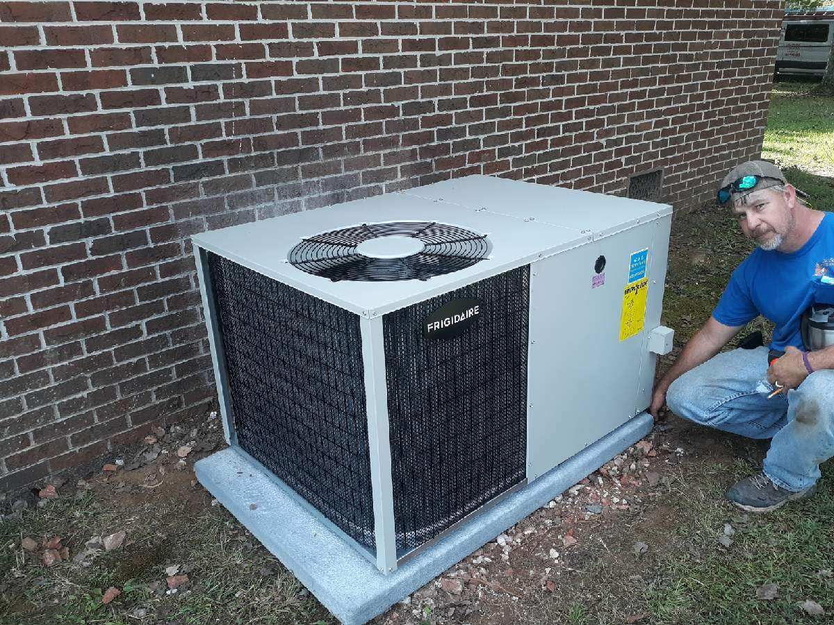 Outdoor HVAC unit beside brick wall, with a worker kneeling nearby inspecting it.