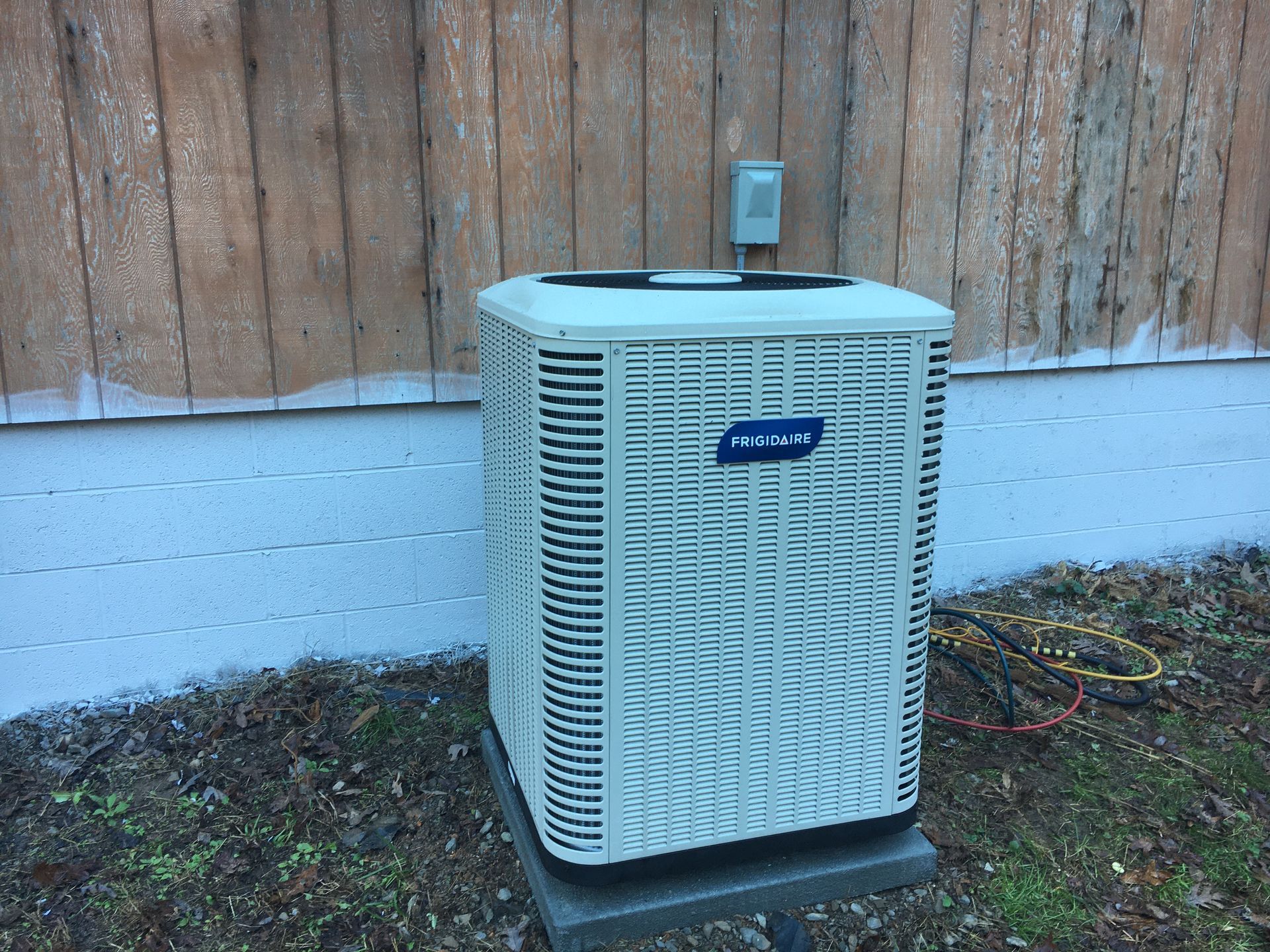 Outdoor air conditioning unit beside a house, with a wooden fence behind it.