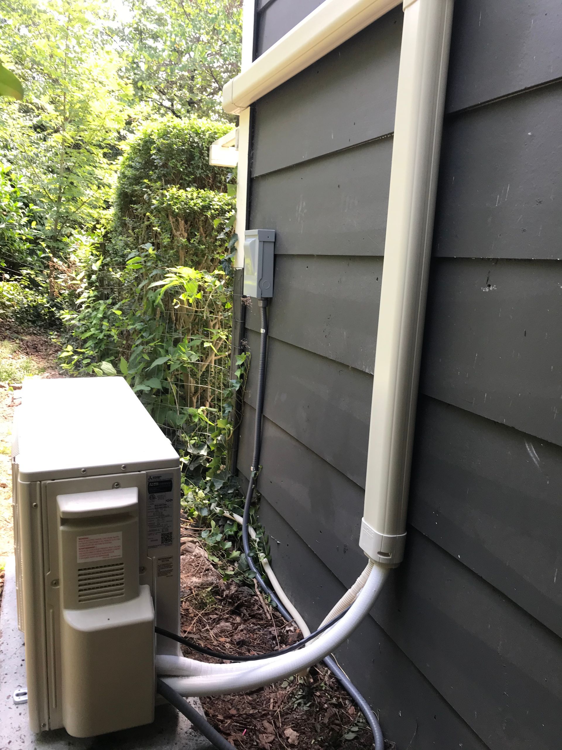 Outdoor air conditioner unit beside dark gray house siding with a white hose and conduit attached
