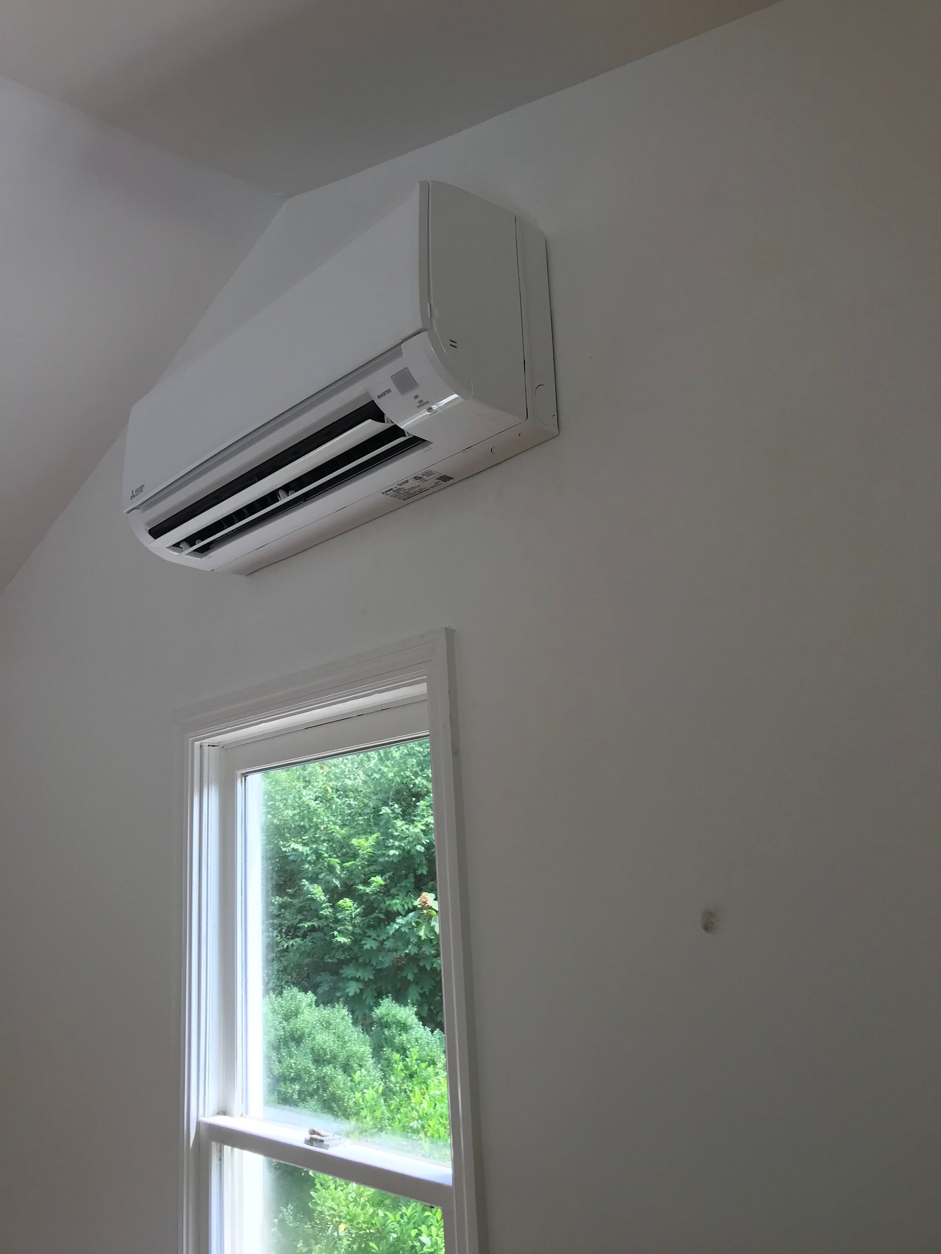 White wall-mounted air conditioner above a window in a bright room with green trees outside.