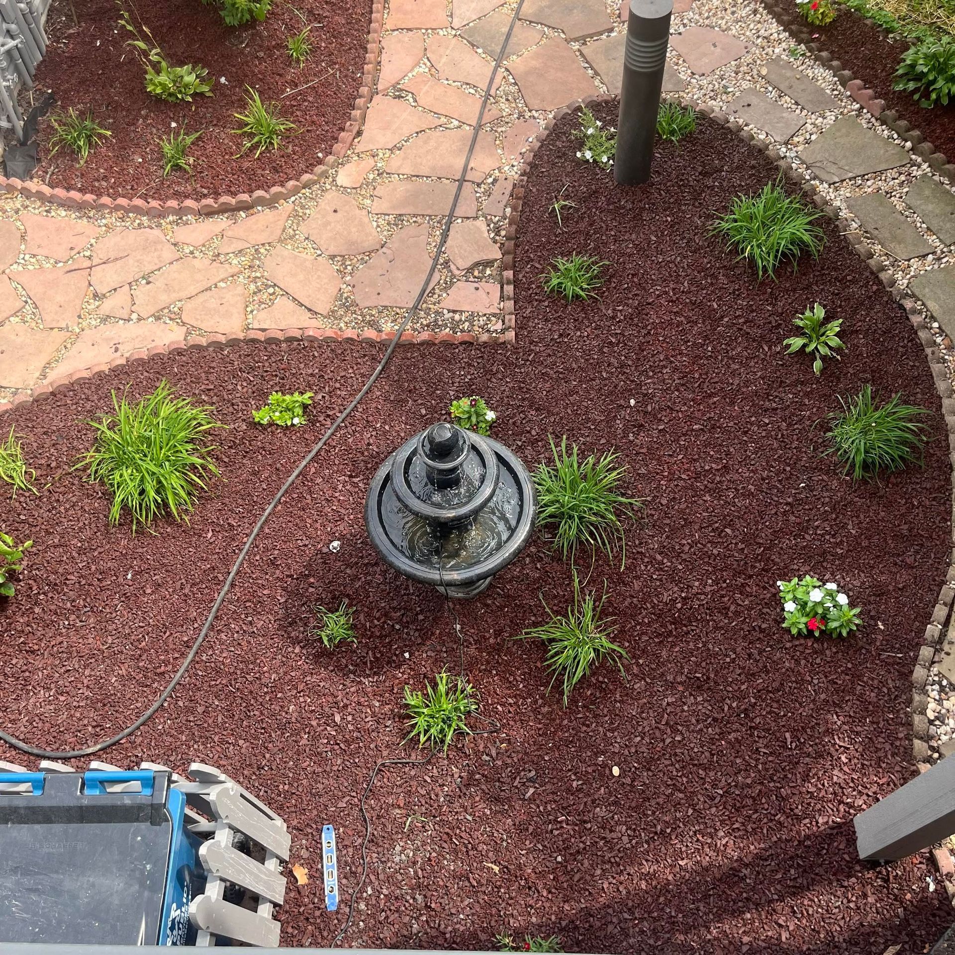 Top-down view of a landscaped garden with a small fountain, red mulch, stepping stones, and scattered plants.