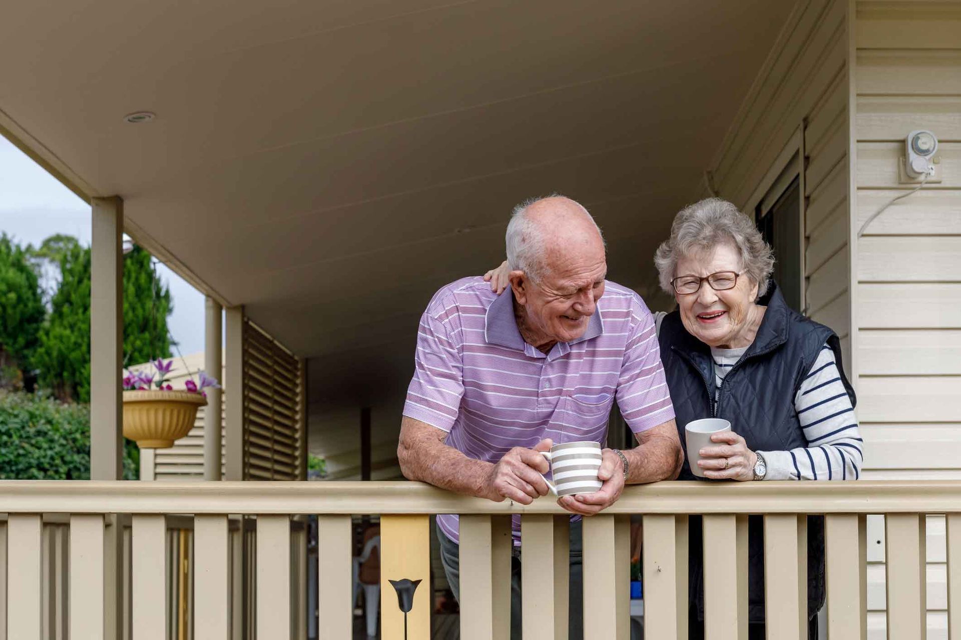 An elderly couple is standing on a porch drinking coffee.