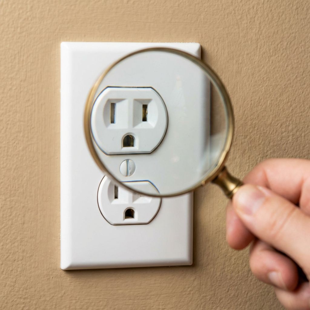 Hand holding a magnifying glass over a white electrical wall outlet on a beige wall