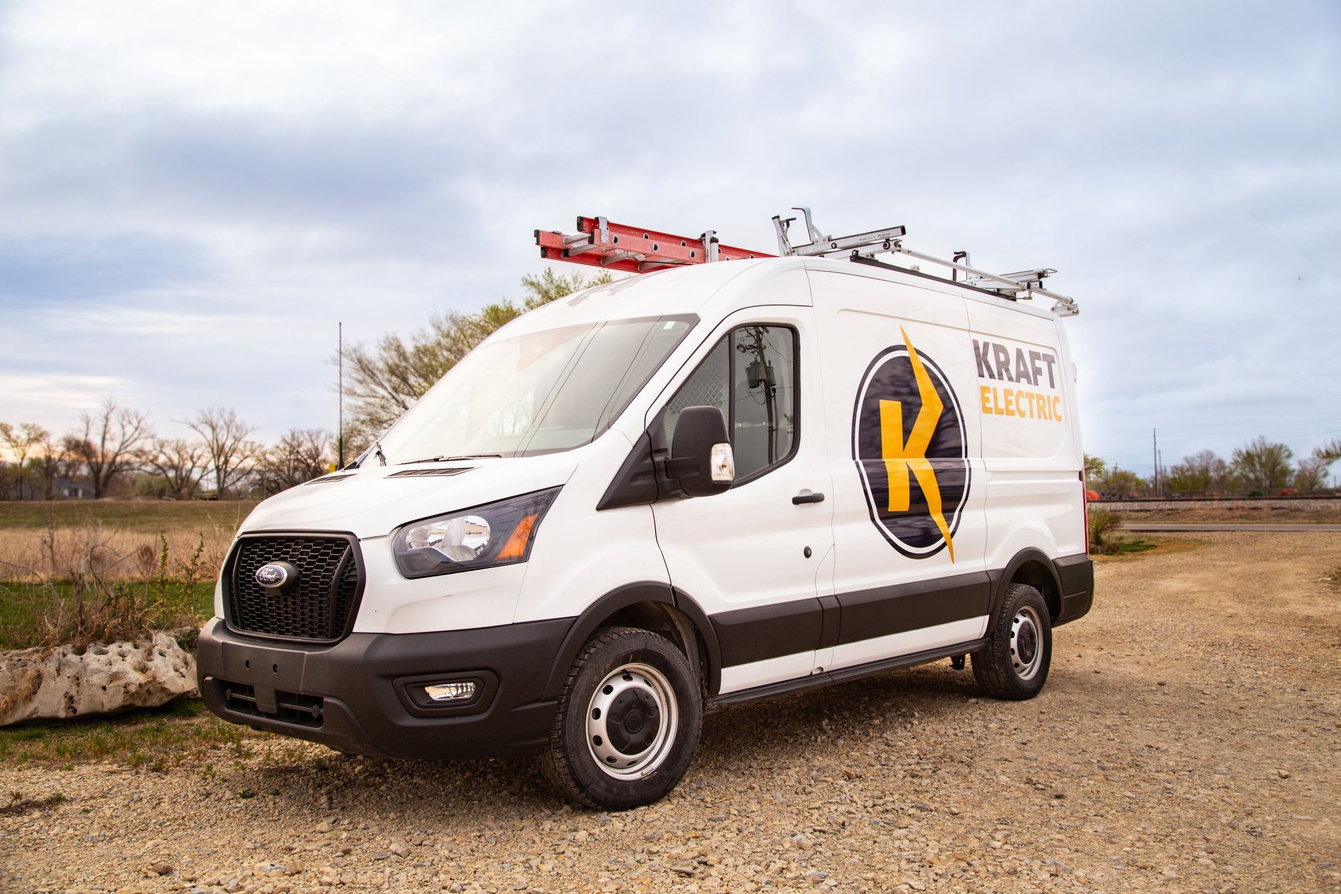 White utility van with ladder rack and large logo parked on a gravel lot under a cloudy sky