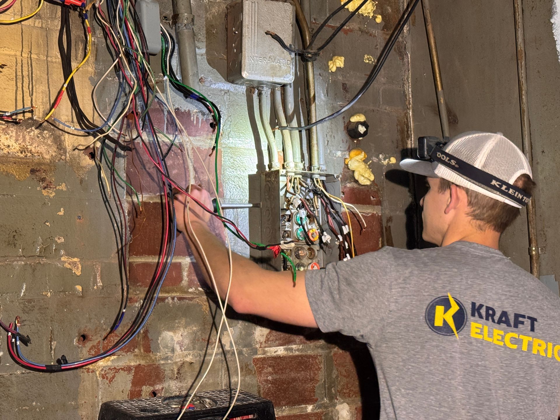 Electrician working on exposed wiring and breaker panels on a brick wall.