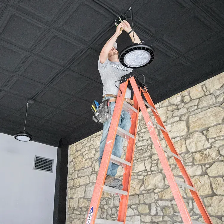 Worker on a red ladder installing a round ceiling light in a room with stone walls and a black ceiling.