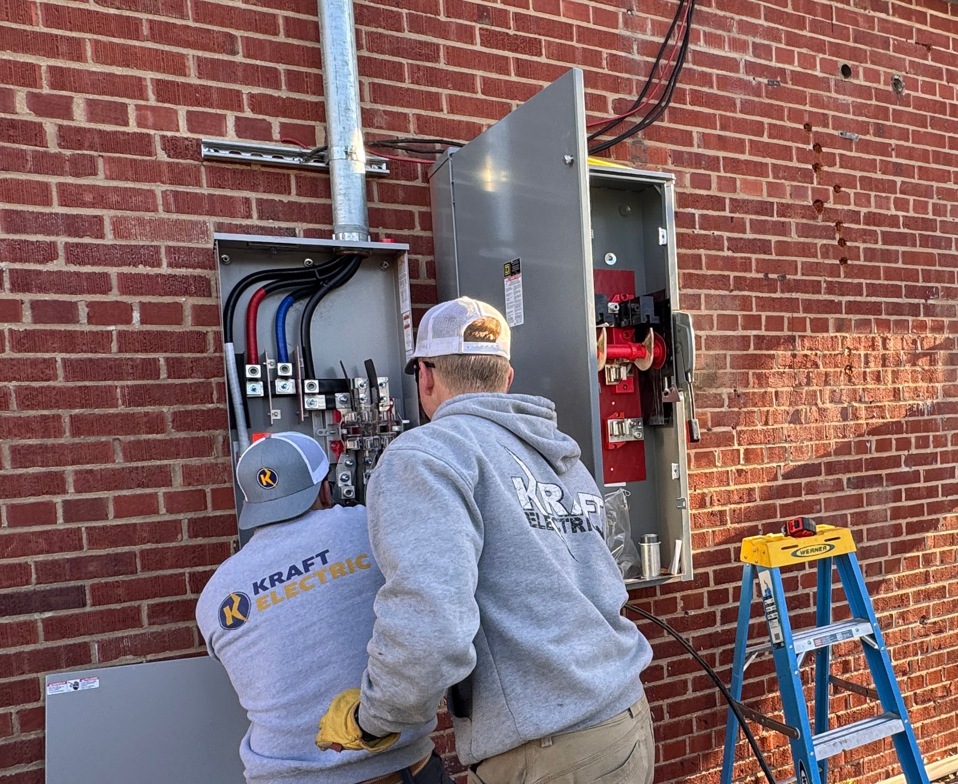 Two workers inspect an open electrical panel on a brick wall, with a ladder beside them.