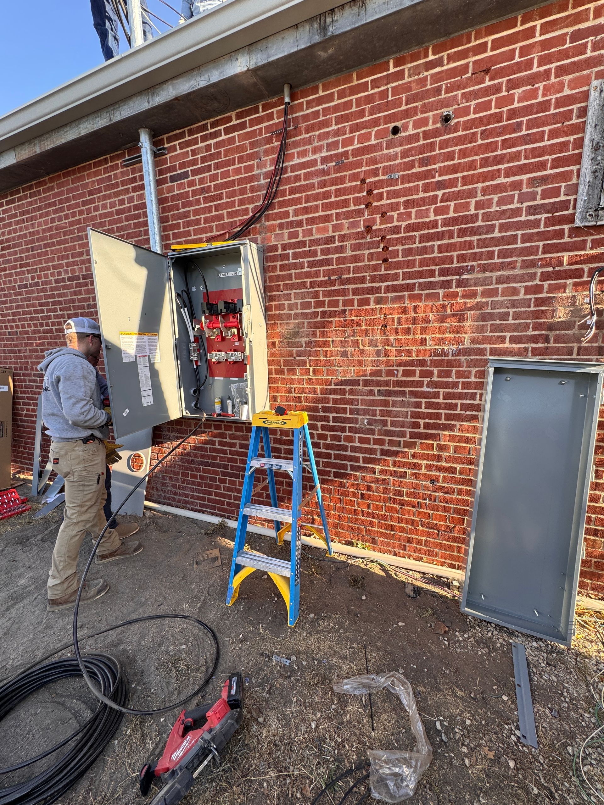 Worker stands by a brick wall with an open utility box and ladder at a construction site.