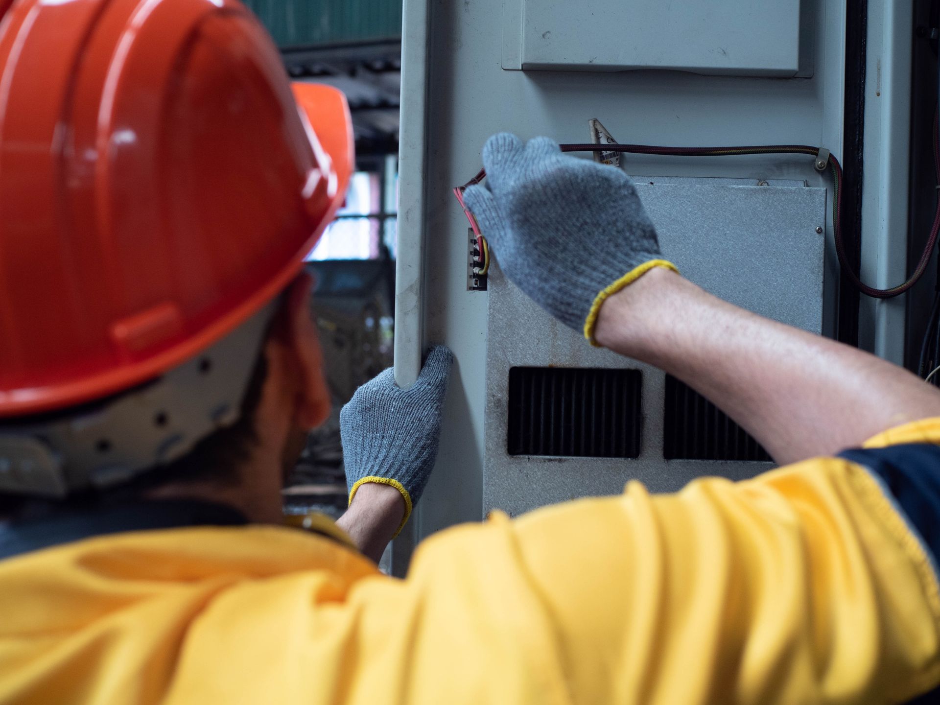 Worker in orange hard hat and yellow shirt inspecting an electrical panel outdoors