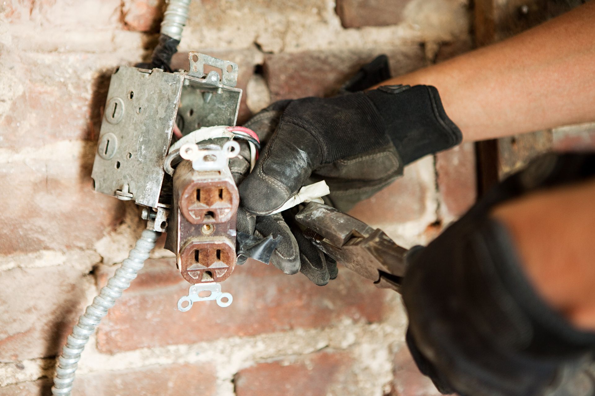 Gloved hand installing an electrical outlet into a wall with exposed wiring and brick background