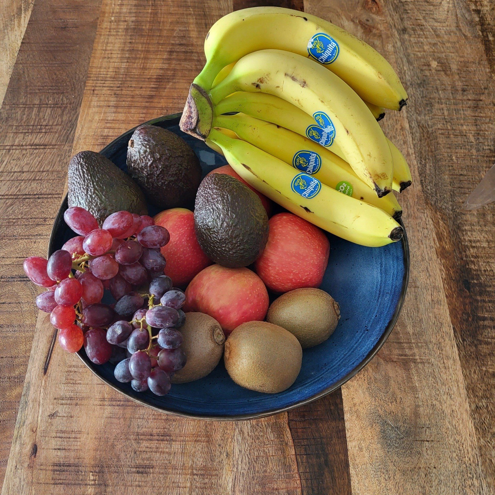Fruitschaal met bananen, druiven, avocado's, appels en kiwi's op een houten tafel.