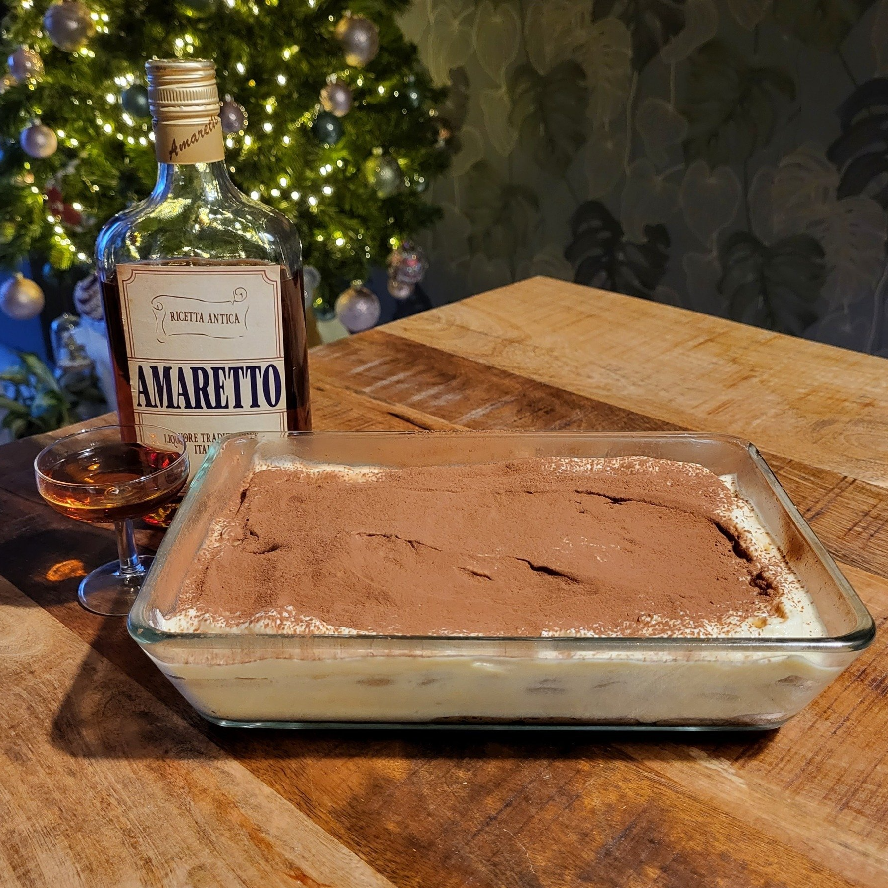 Tiramisu-dessert met cacaopoeder, naast een fles Amaretto en een cocktailglas op een houten tafel.