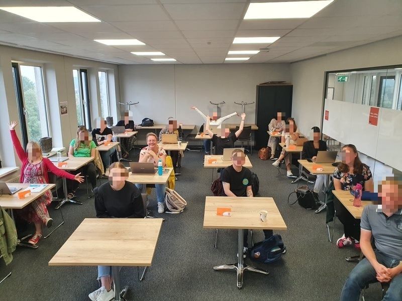 Een groep mensen in een klaslokaal, sommigen met opgeheven handen. Er zijn tafels en laptops aanwezig.