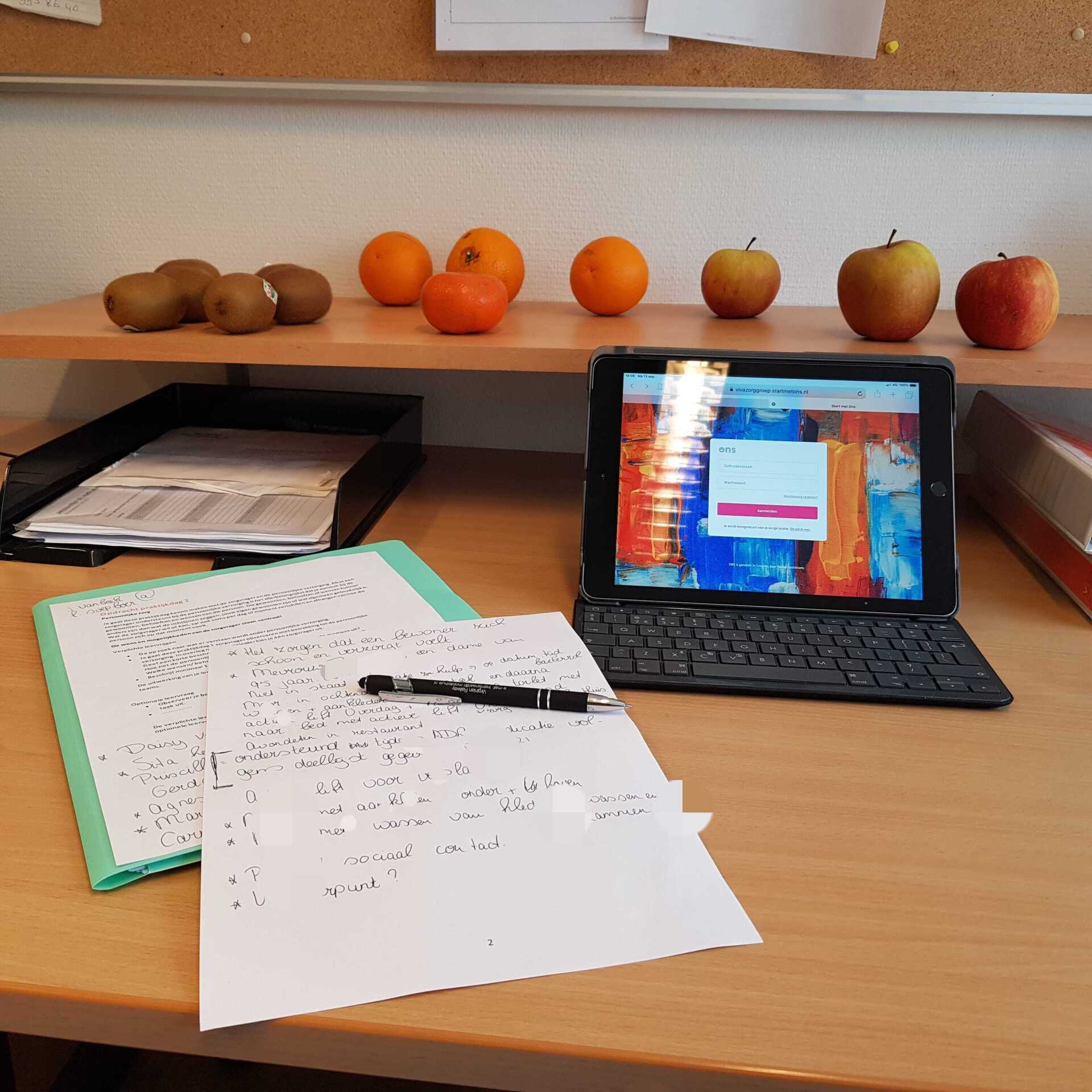 Een bureau met fruit (kiwi's, sinaasappels, appels), een tablet, papier en een pen in een goed verlichte ruimte.