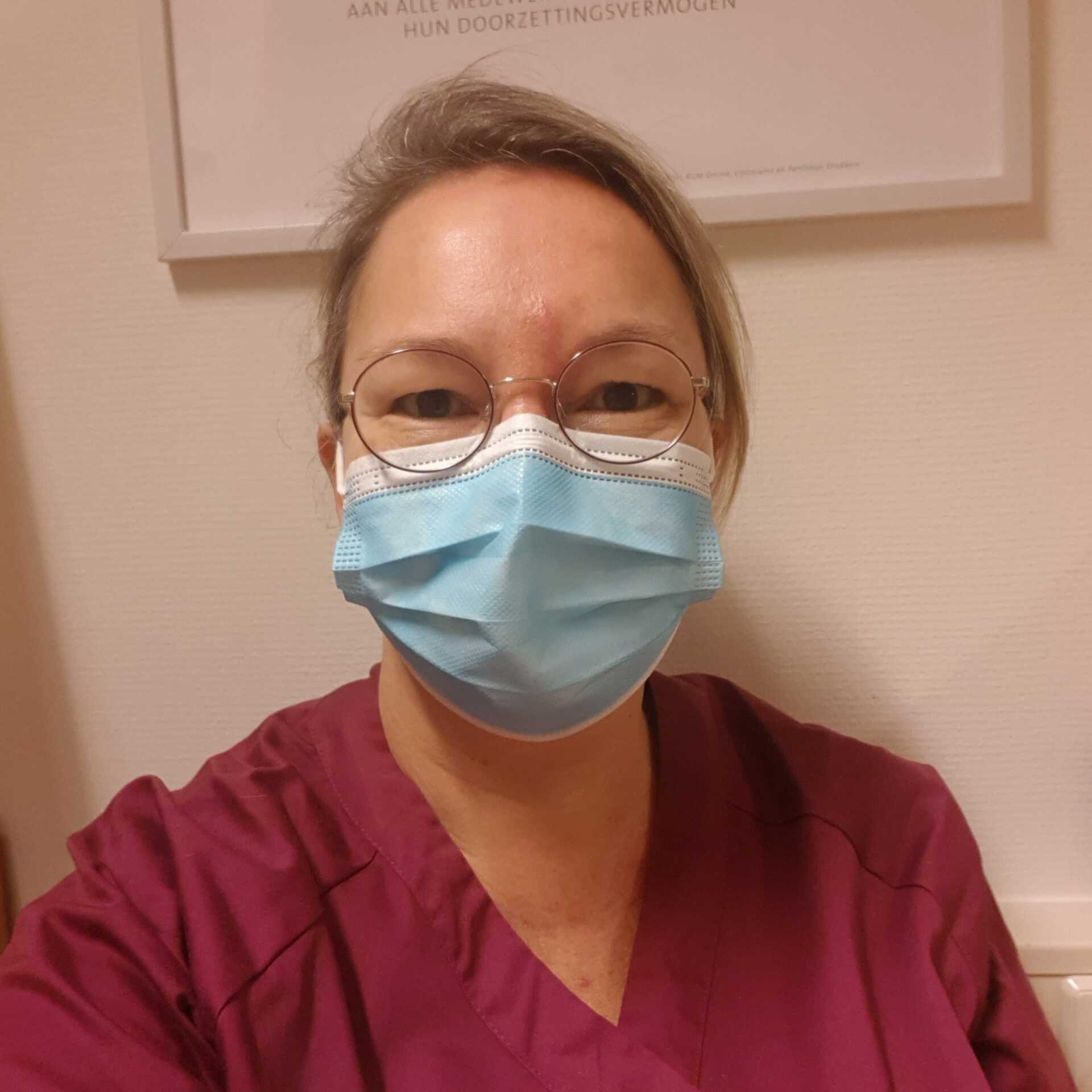 Vrouw met een gezichtsmasker, een bril en kastanjebruine kleding, binnenshuis.