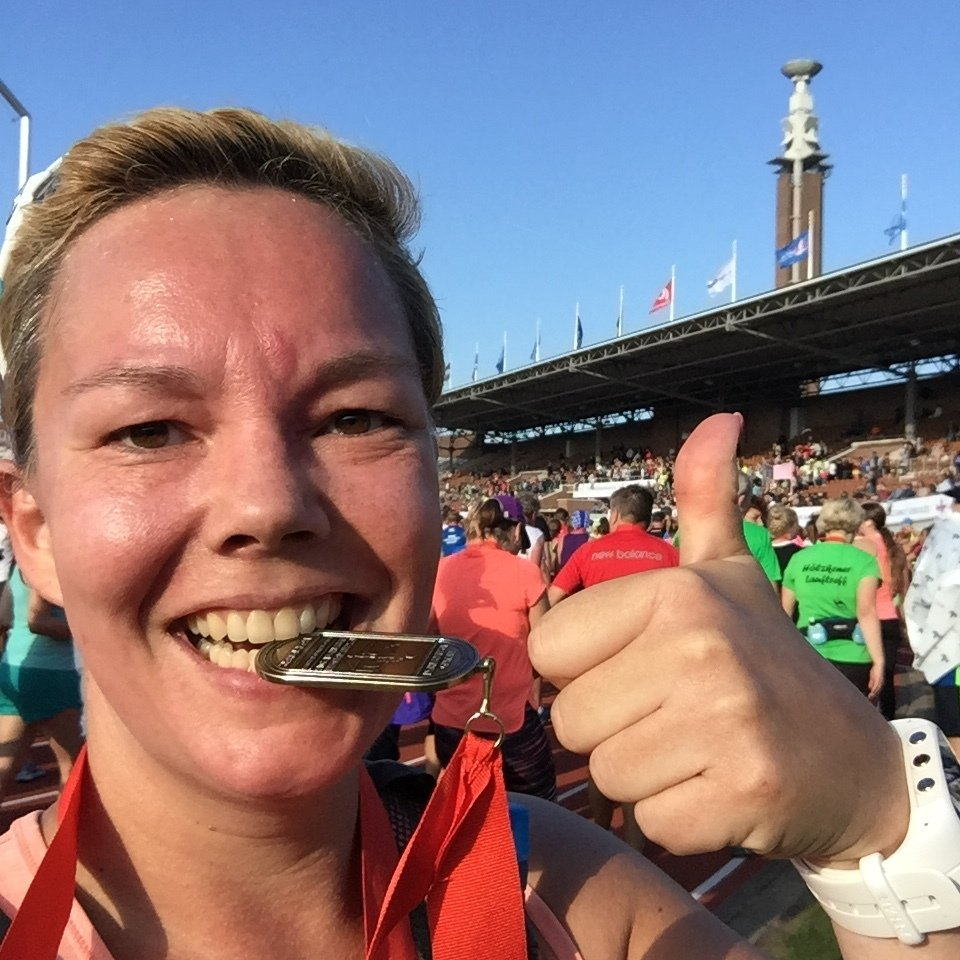 Vrouw met medaille, lachend, duim omhoog, voor het stadion, in hardloopkleding.