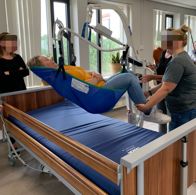 Vrouw wordt met een tillift in bed getild, geholpen door twee mensen. Anderen kijken toe. Blauwe draagdoek, binnen.