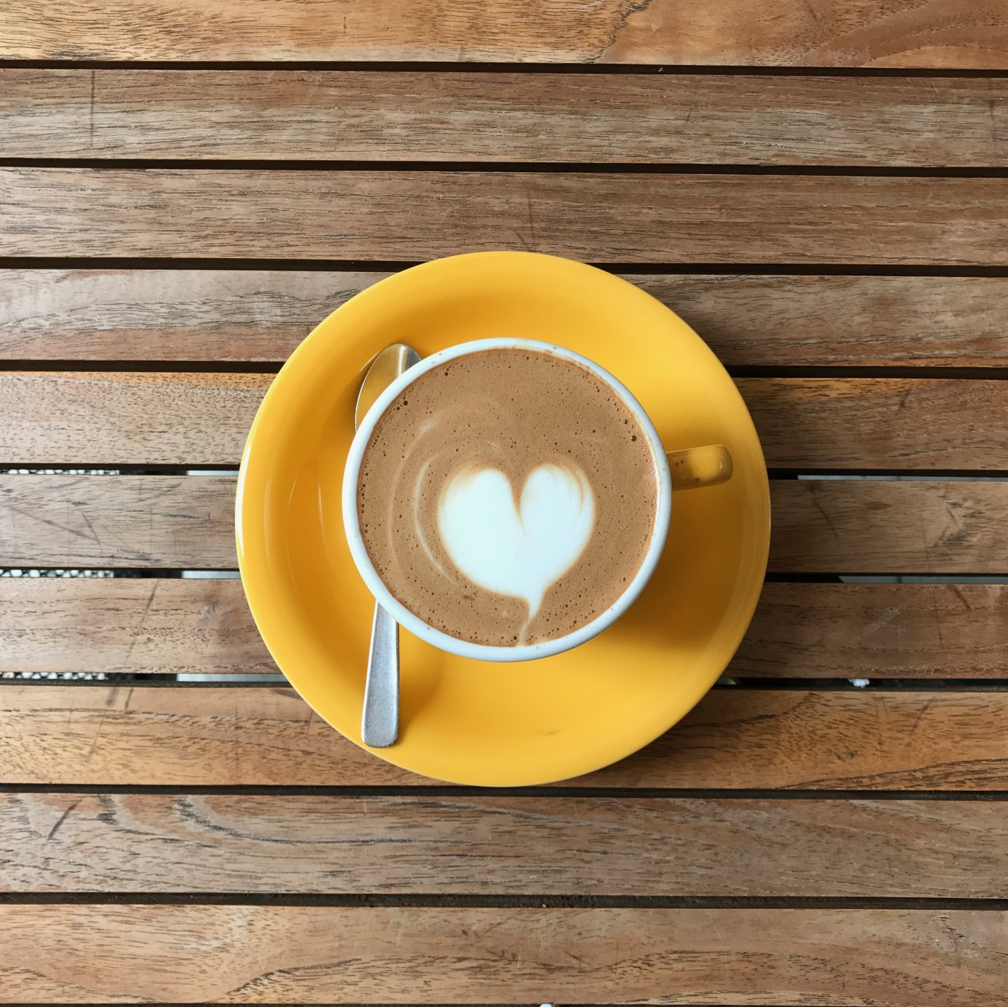 Gele kop koffie met hartvormig schuimkunstwerk op een geel schoteltje met een lepel op een houten tafel.