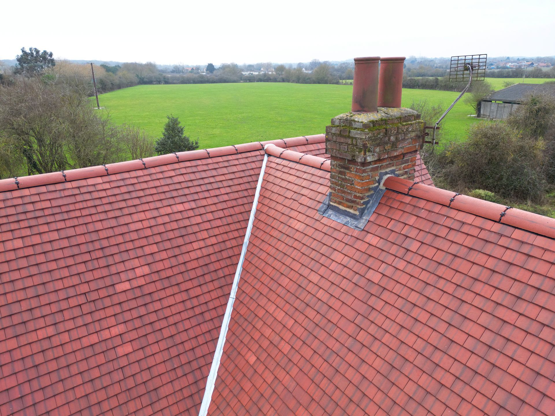 An aerial view of a brick chimney with clay flue pots on a red tile roof above a green field