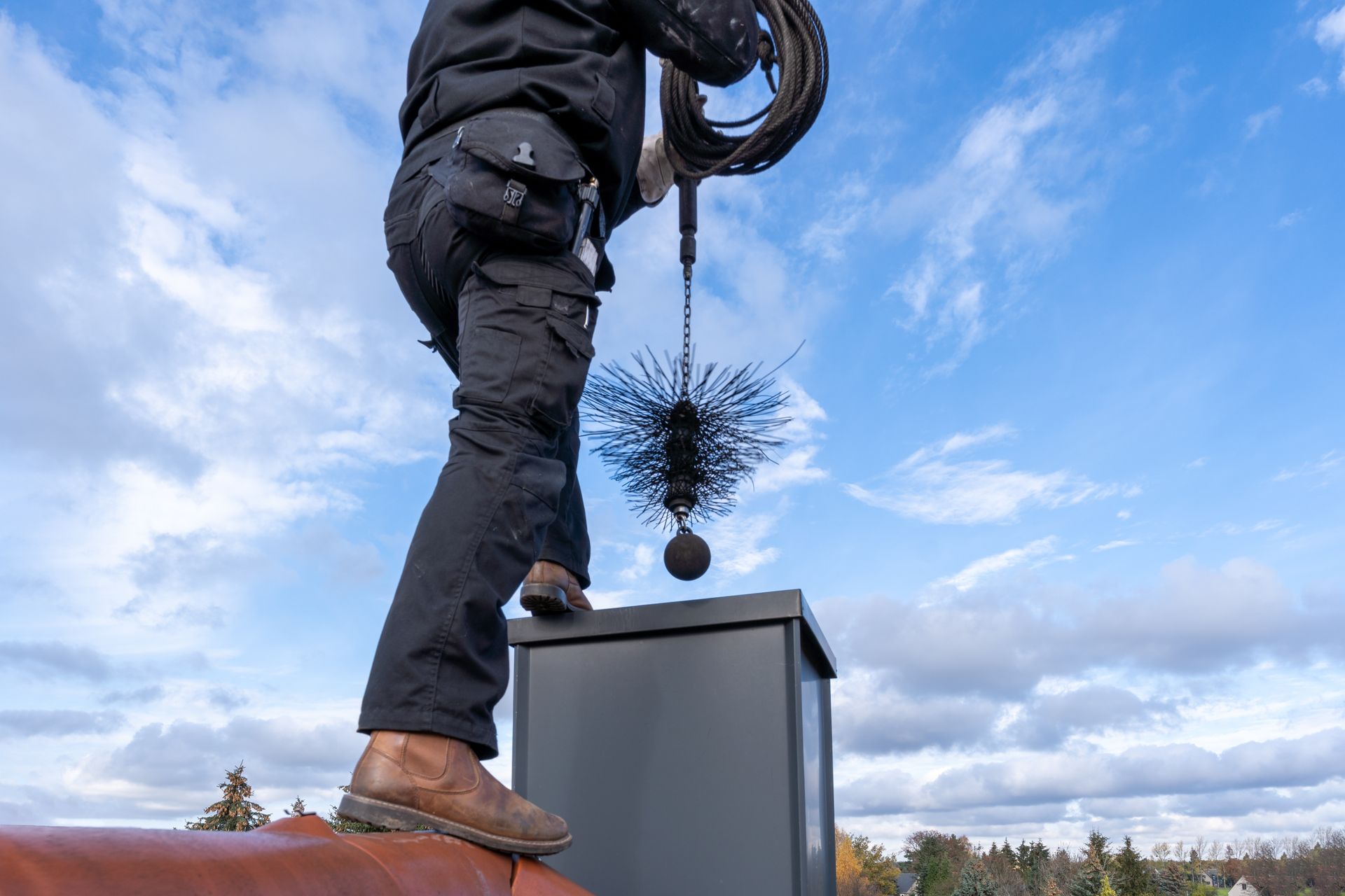 A chimney sweep cleaning a chimney standing on the house roof, lowering equipment down the flue