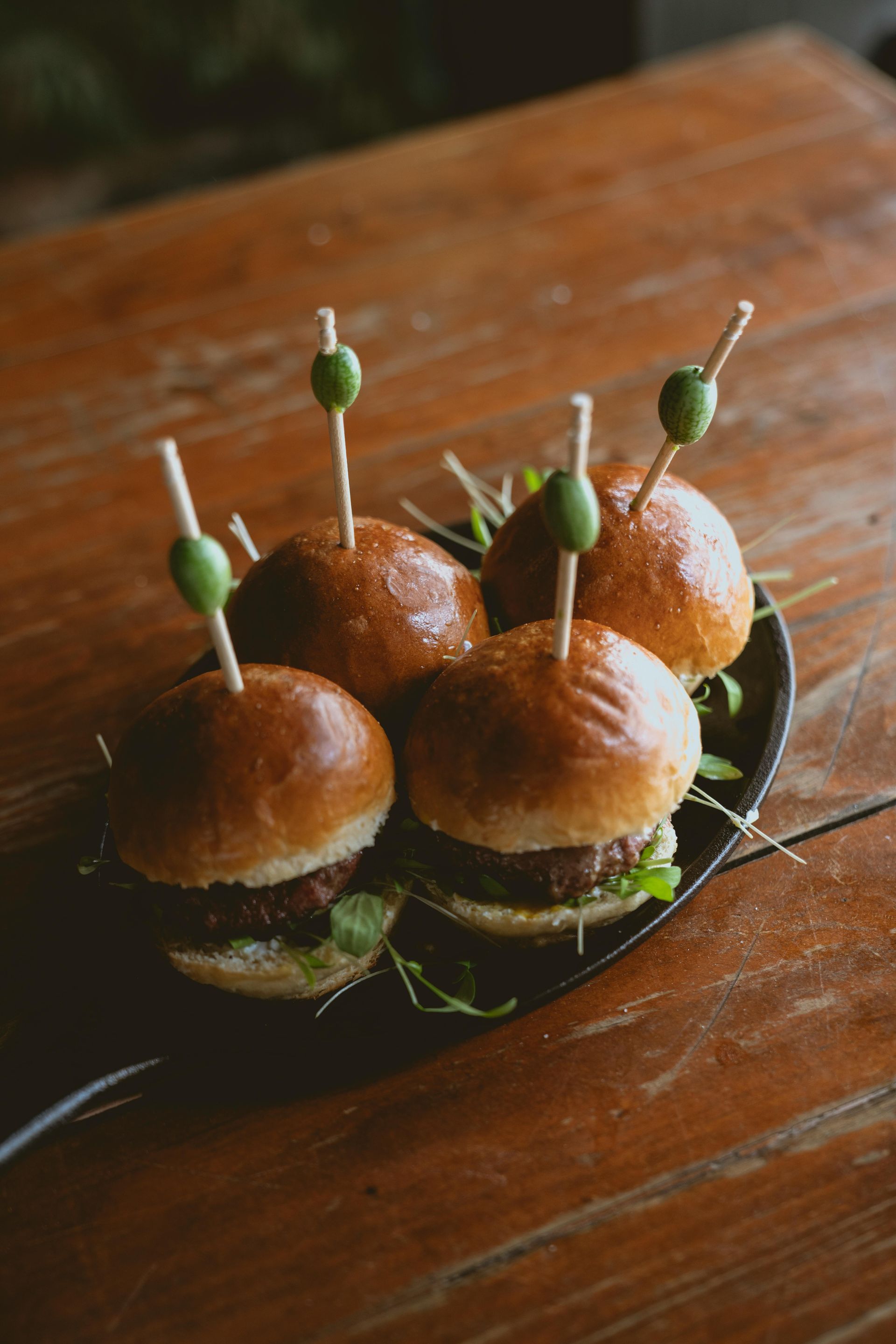Mini burgers on a black platter, skewered with olives, on a wooden table.