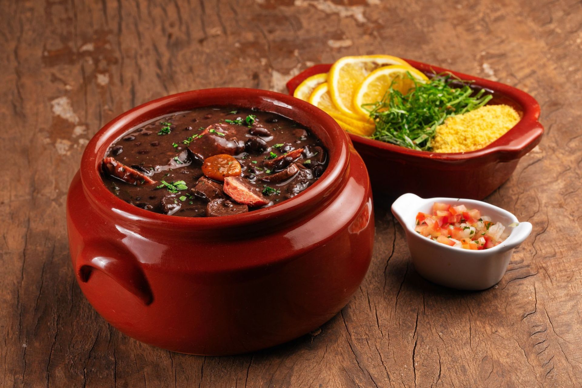 Feijoada, Brazilian black bean stew, served in a ceramic pot with side dishes on a wooden table.