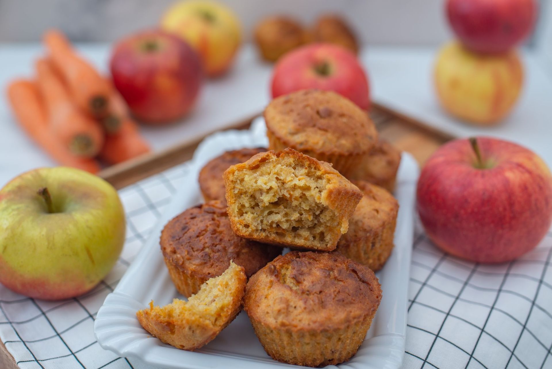 Muffins on a tray, surrounded by apples and carrots on a checkered cloth. One muffin is cut, showing the inside.