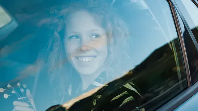 Mulher sorridente vista através da janela de um carro, com reflexos do céu azul.