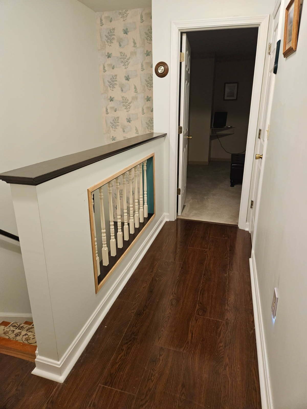 Hallway with dark wood floor, white walls, and a staircase railing. A door leads to another room.