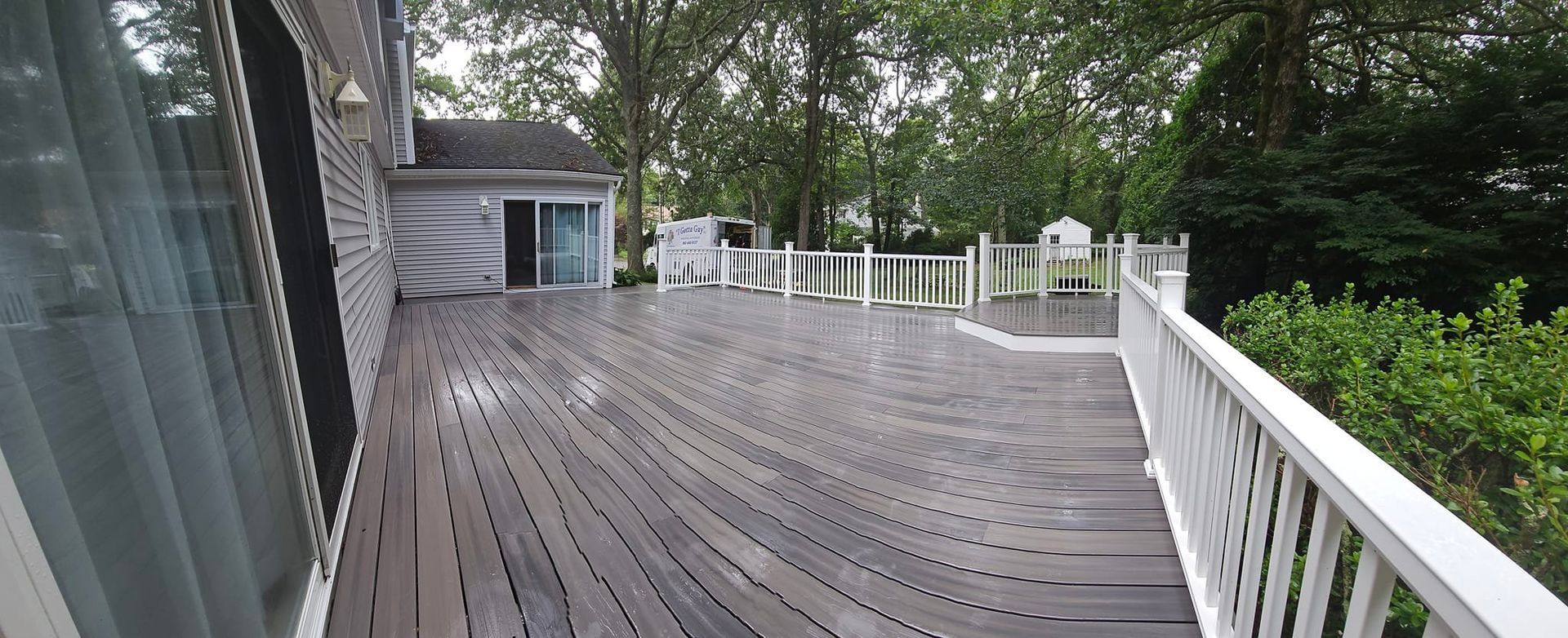 A wooden deck with white railing around it. Trees and a house in the background.