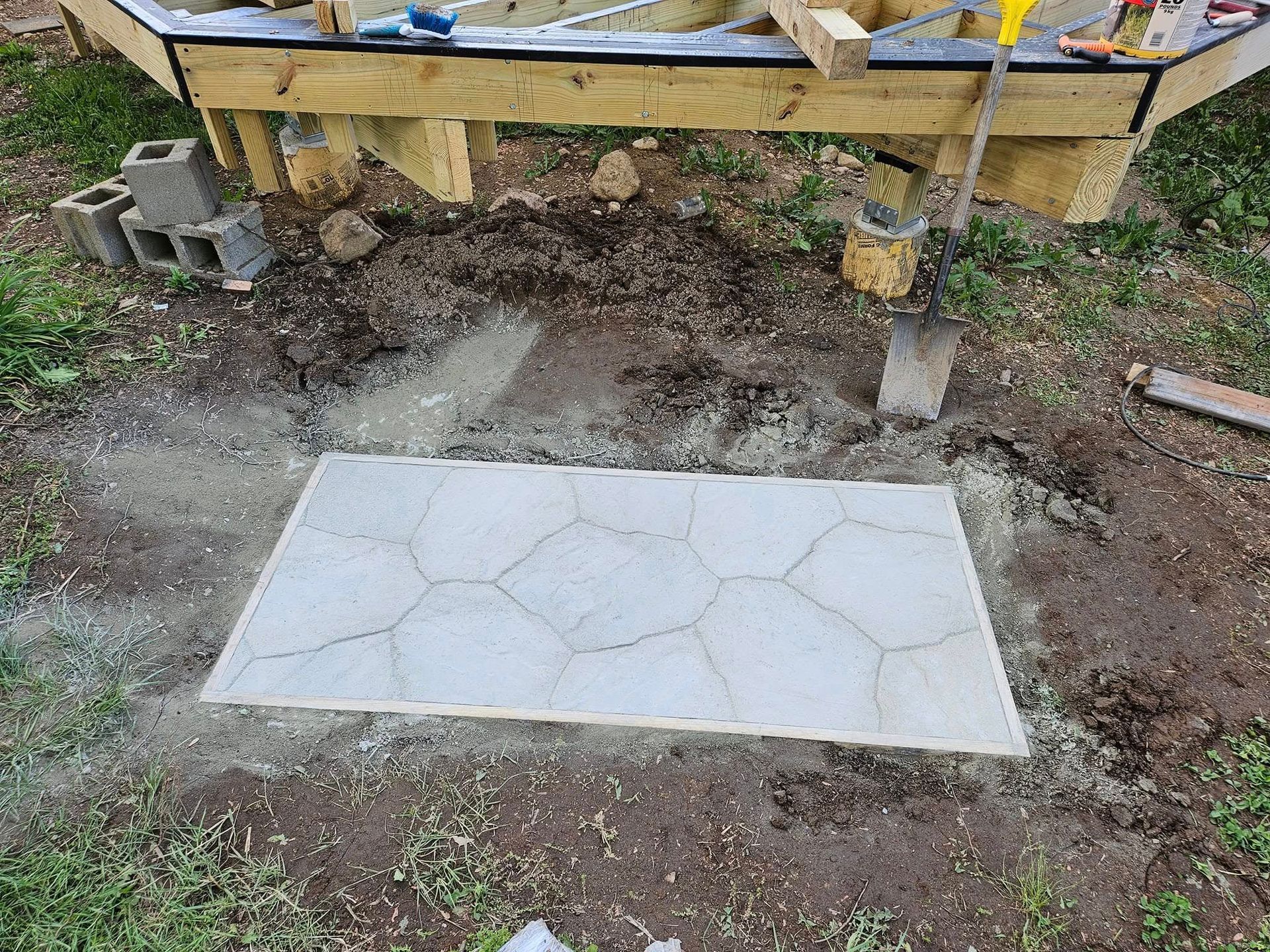A newly poured, light gray concrete patio with a cracked pattern sits under a wooden deck under construction.