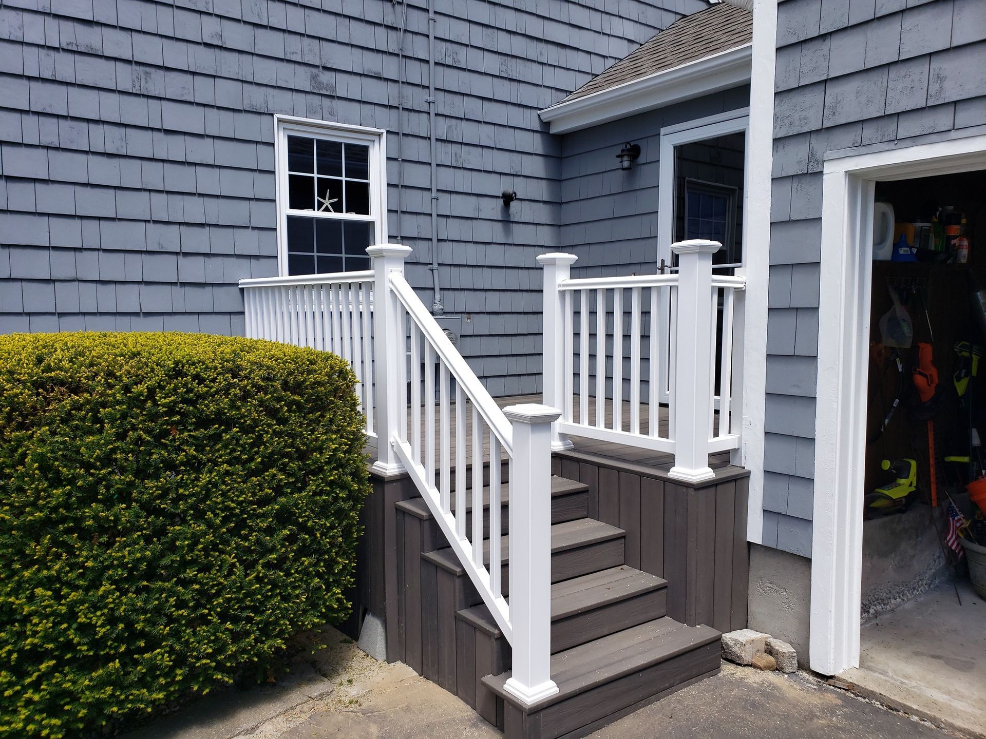Gray house with white railing and steps leading up to the door and a green bush.