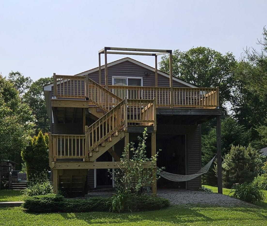 Two-story house with wooden decks and stairs. Hammock hangs below the upper deck. Green foliage surrounds the house.