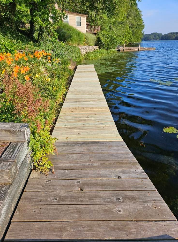 Wooden dock extending into a lake, flanked by flowers and foliage. Blue water and sky.