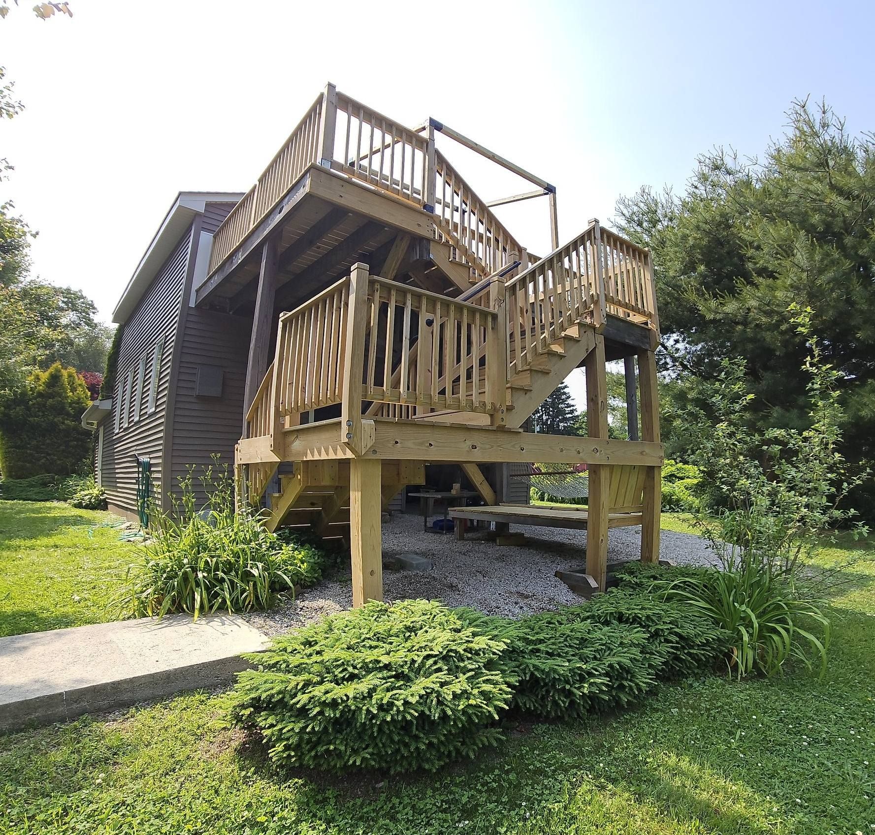 Wooden deck with stairs attached to a two-story building surrounded by greenery and a stone path.
