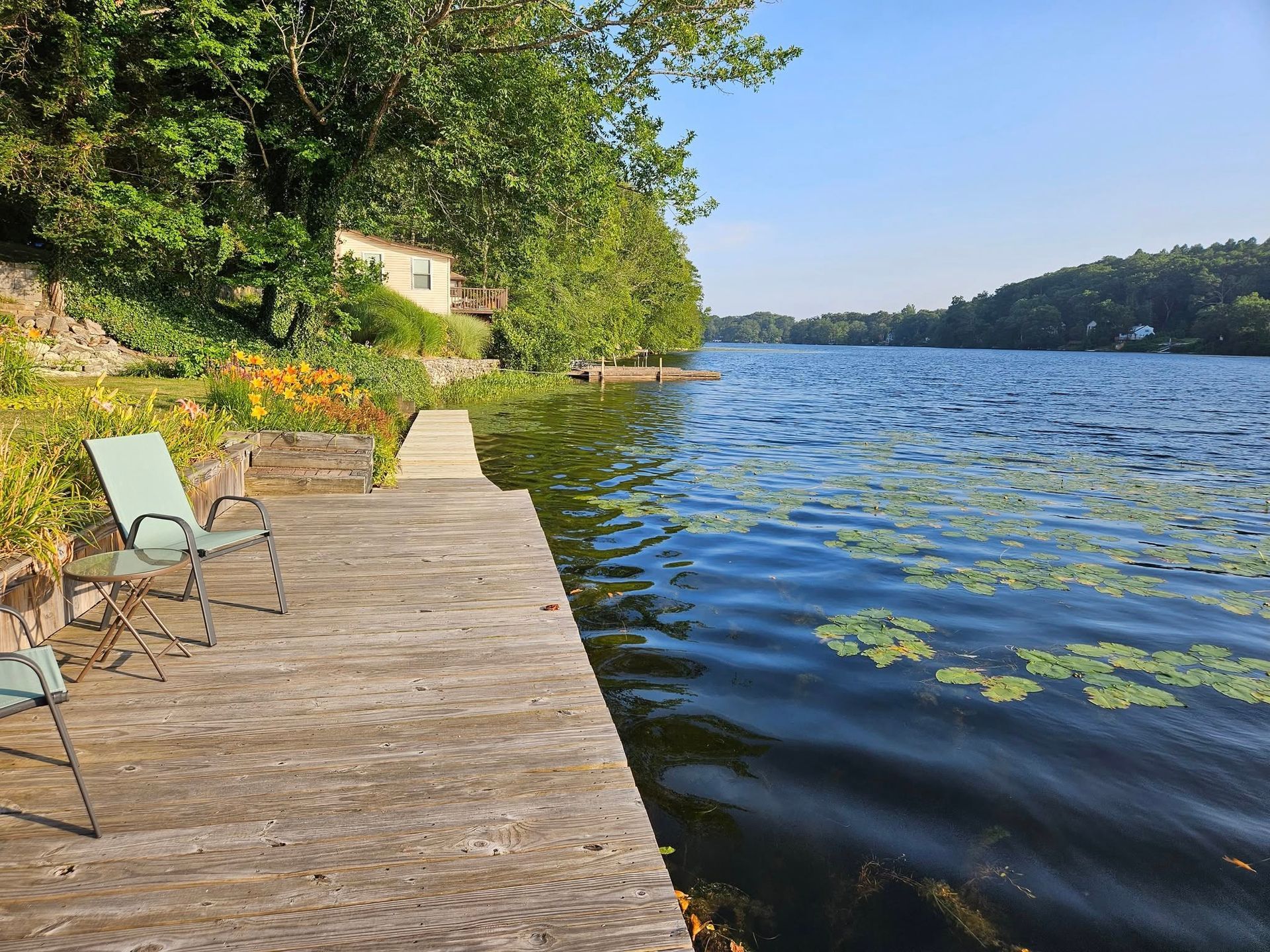Lakefront scene: weathered dock with two green chairs, water, trees, and flowers under a sunny blue sky.