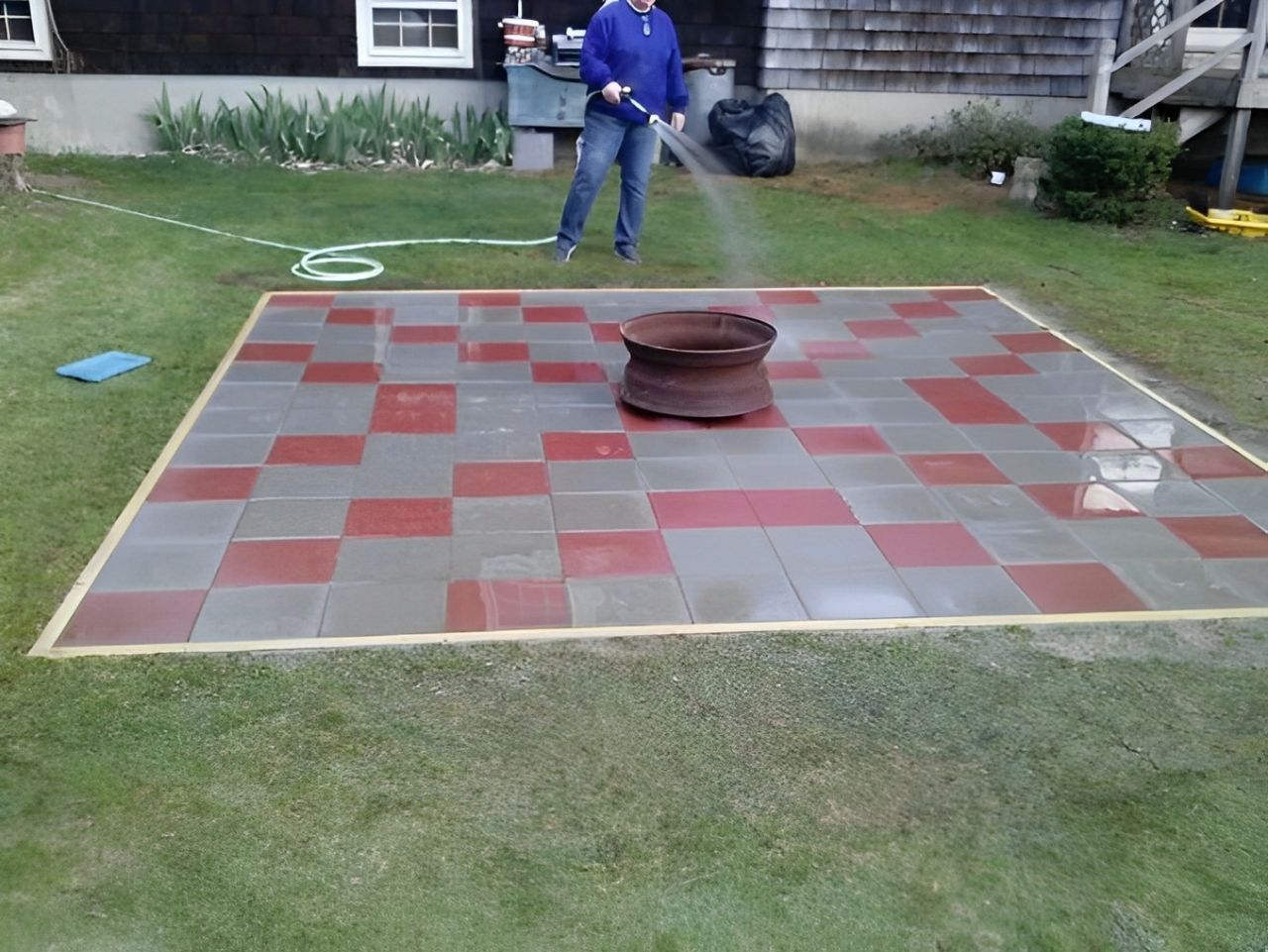 Person spraying a red and gray tiled patio with a hose, fire pit in the center.