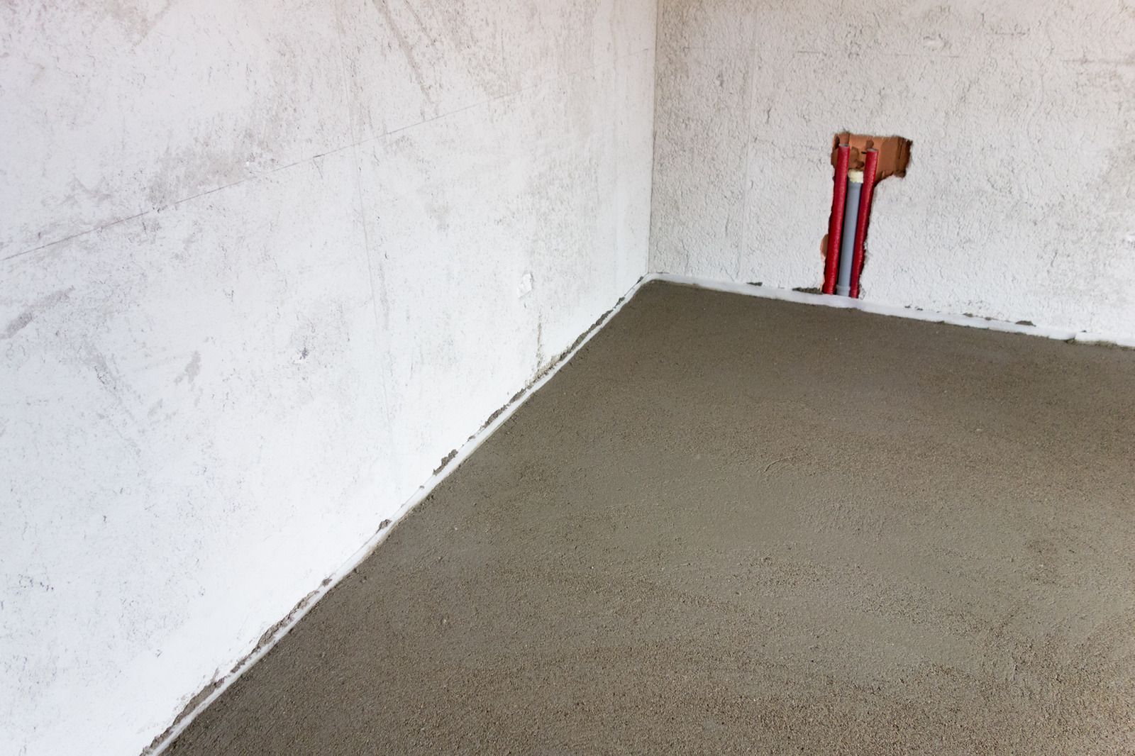 Freshly poured concrete floor in a building corner, next to white walls, with red pipes emerging.