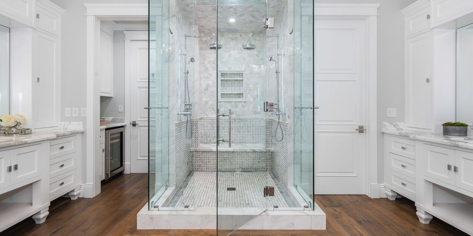Glass shower enclosure in a bright white bathroom with double vanities and marble flooring