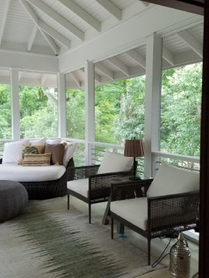 Covered white porch with wicker seating and a round daybed overlooking lush green trees.