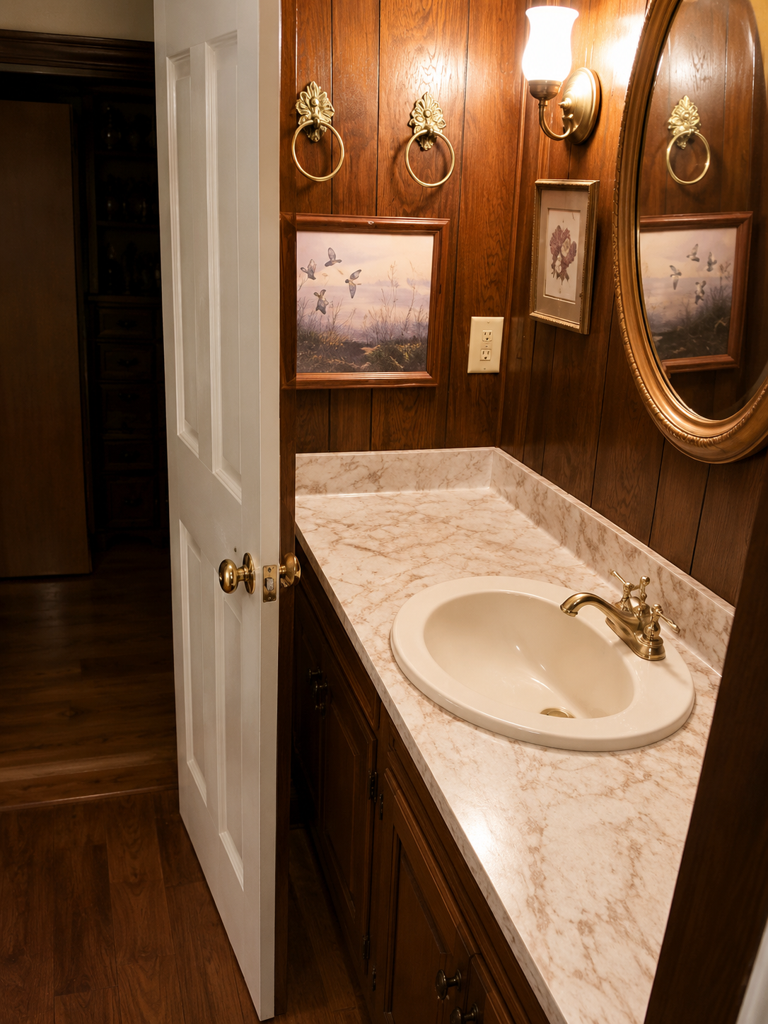 Small bathroom vanity with cream sink, marble countertop, wood paneling, and decorative mirrors and framed art