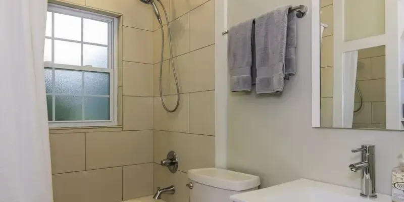 Small beige bathroom with window, toilet, shower, sink, mirror, and towels hanging on the wall
