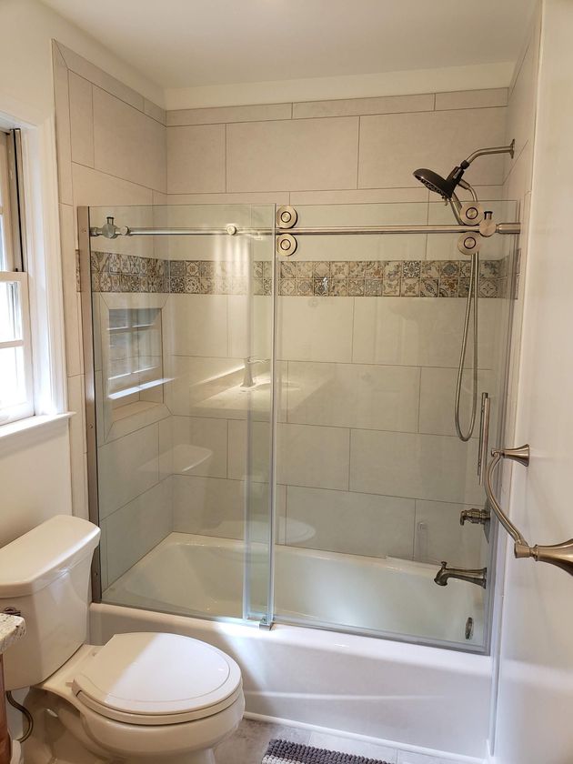Bathroom with toilet and glass-enclosed shower over a tub, white tile, and a window on the left.